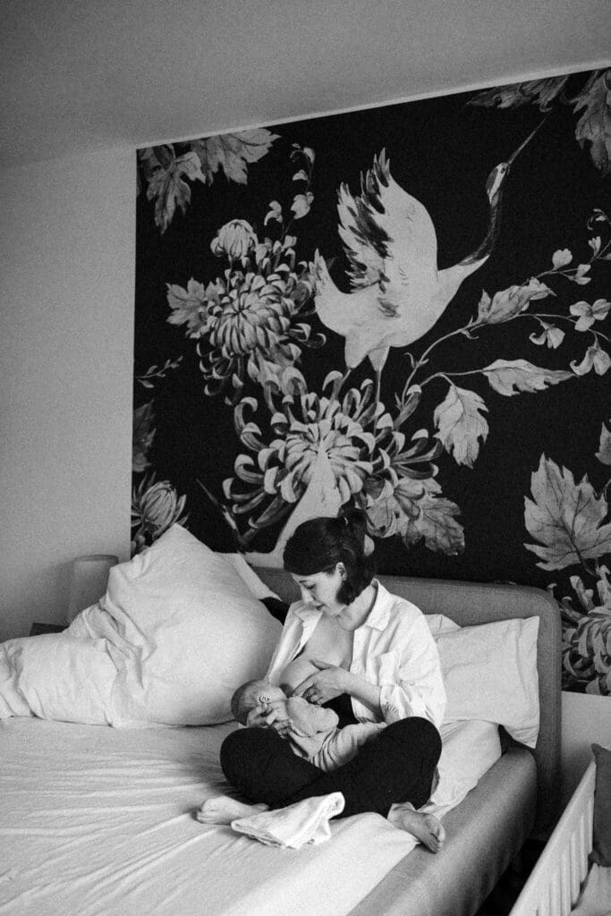 Mother breastfeeds her newborn on the bed in front of crane-and-floral wallpaper during an in-home newborn session in Munich