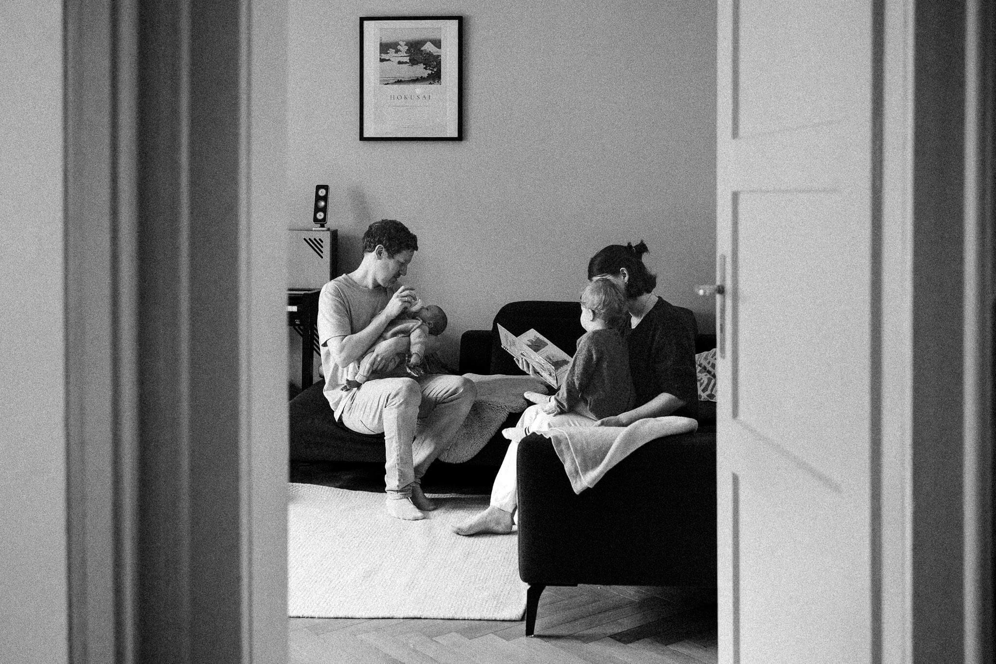 Father holds his newborn while mother reads to their toddler on the couch — a candid family moment from a Munich in-home newborn session