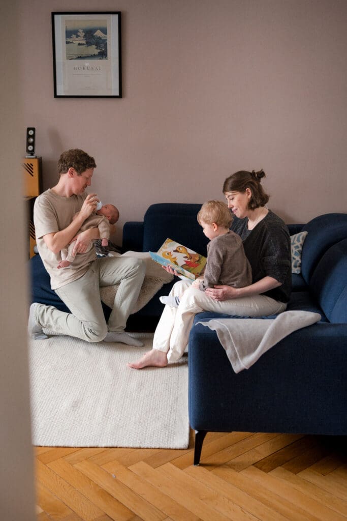 Dad kneels to bottle-feed his newborn while mom reads a picture book to their toddler — lifestyle newborn photography in Munich