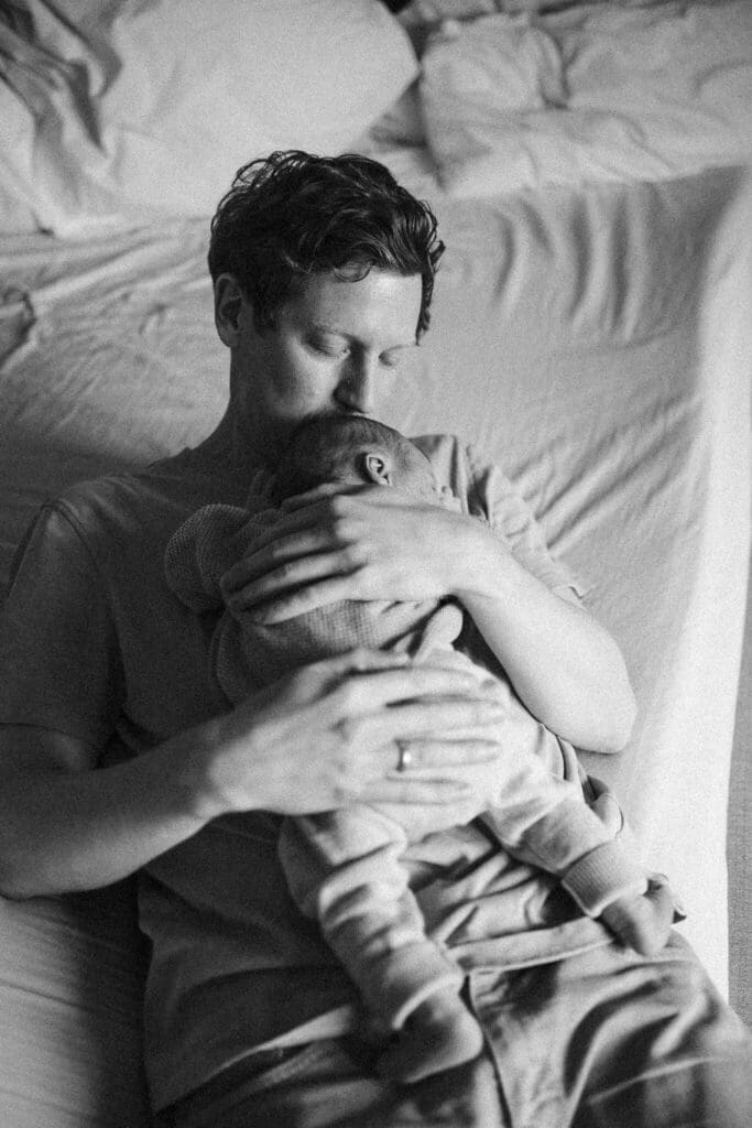 Father presses a quiet kiss to his newborn's head while resting on the bed — captured by a Munich newborn photographer