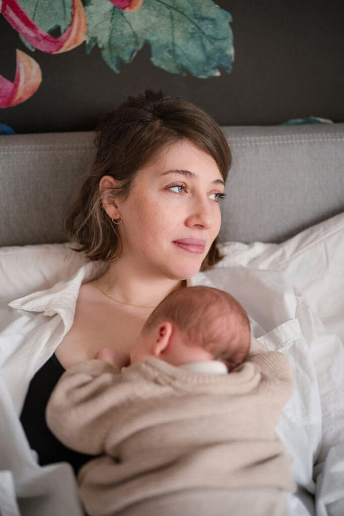 Mother rests in bed cradling her sleeping newborn — an unposed portrait by a Munich newborn photographer.