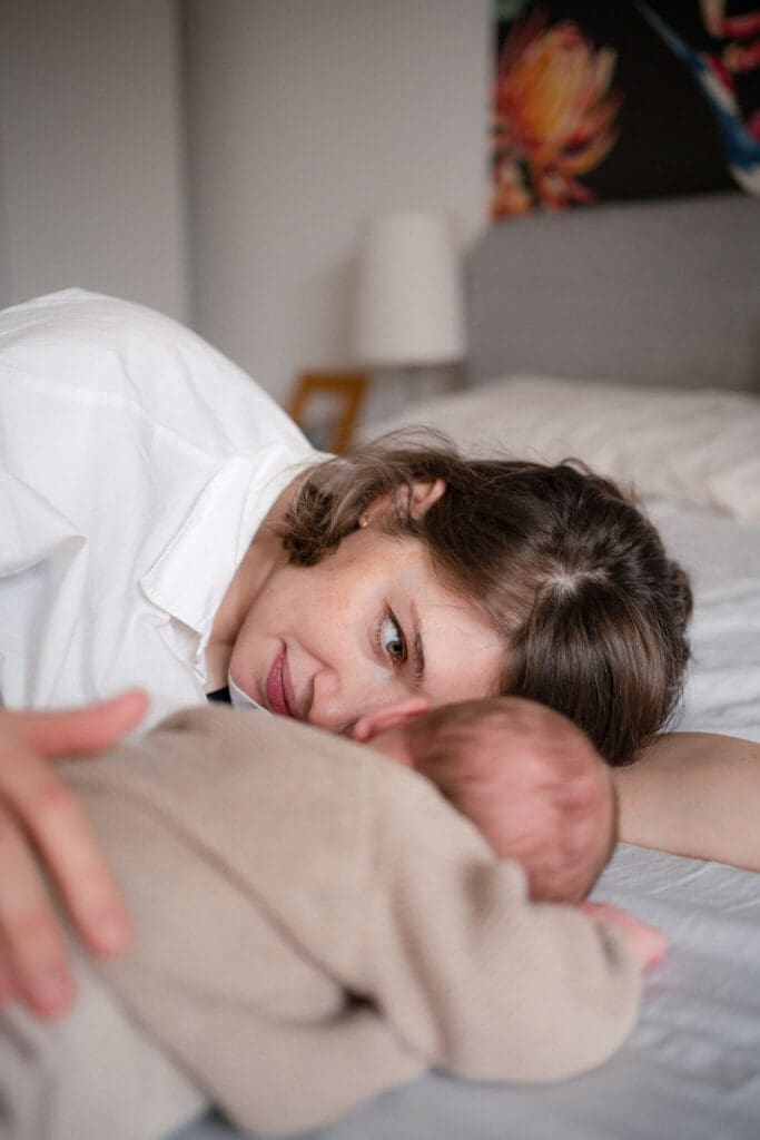 Mother lies on the bed beside her newborn, glancing softly toward the camera — authentic newborn photography in a Munich home