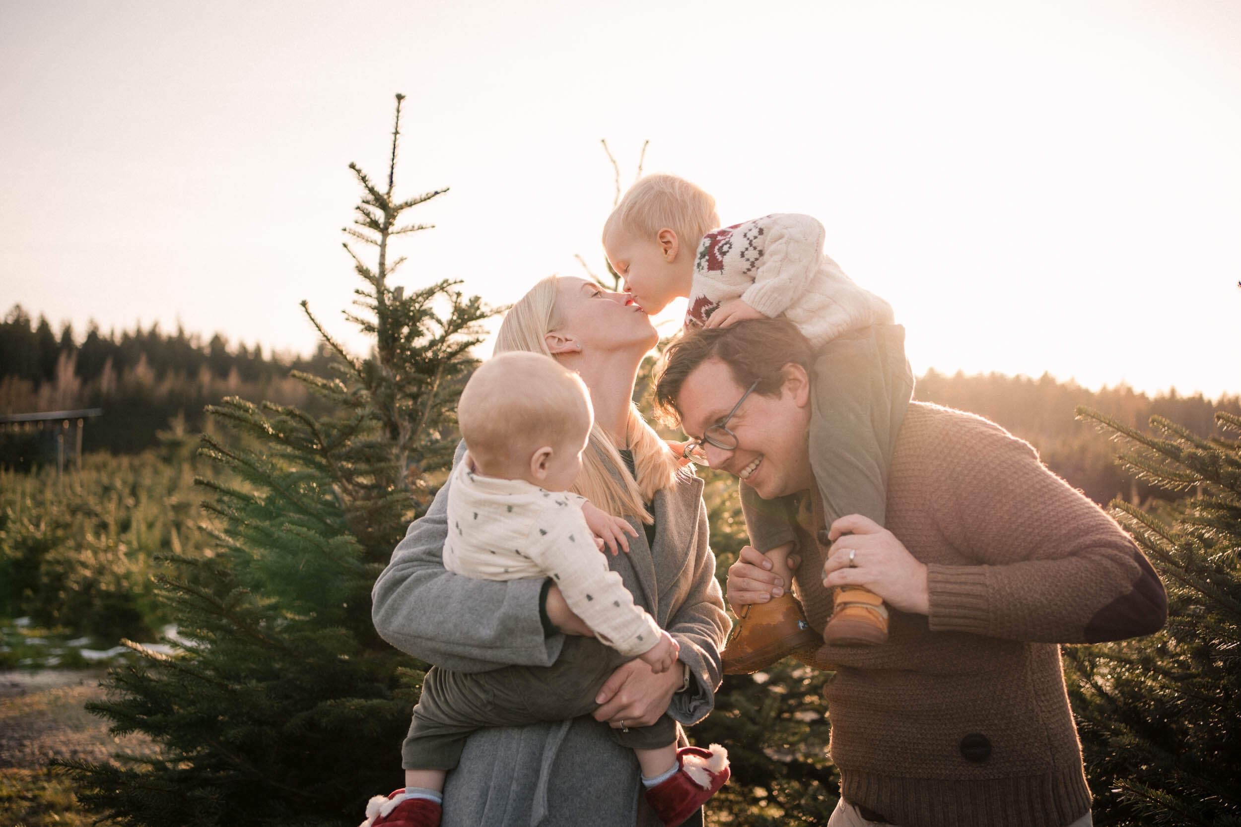 Mom kissing her toddler on dad’s shoulders while holding the baby at sunset during annual family photos in Munich