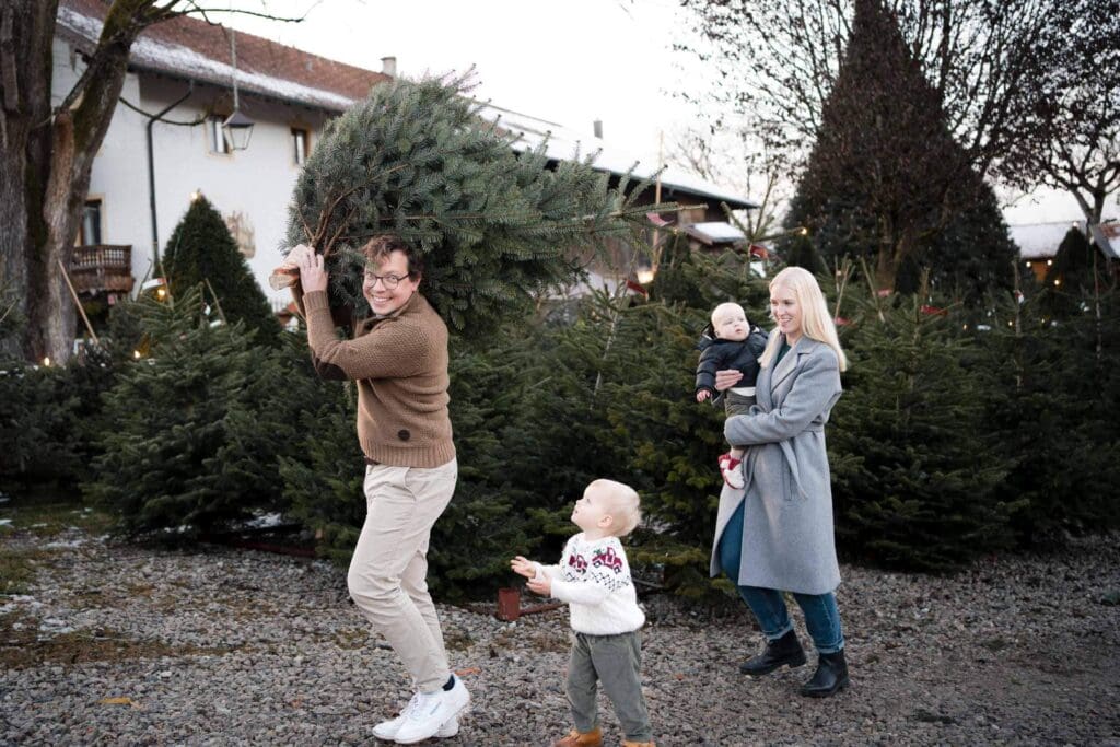 Dad carrying a freshly cut Christmas tree over his shoulder while his family watches at a tree farm near Munich