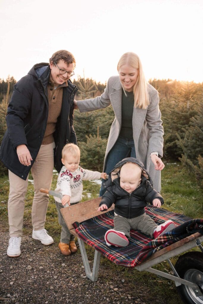 Parents smiling down at their toddler and baby sitting in a wheelbarrow with a plaid blanket at a Christmas tree farm
