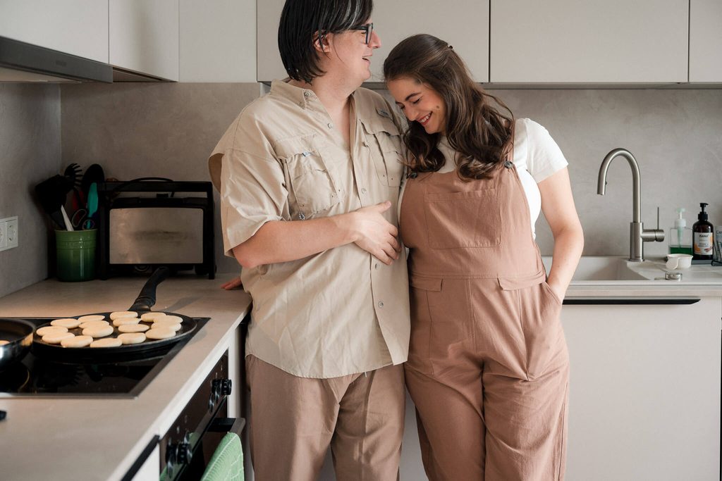 Expecting couple leaning on each other in their kitchen while making breakfast during maternity photos in Munich