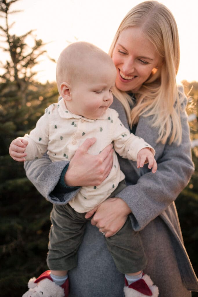 Mom laughing as she holds her baby among the Christmas trees