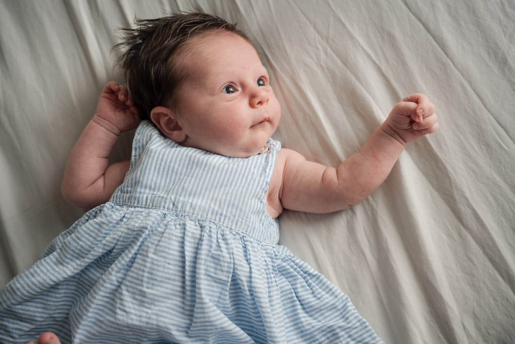 Wide-eyed newborn baby girl in a blue striped dress lying on her parents’ bed