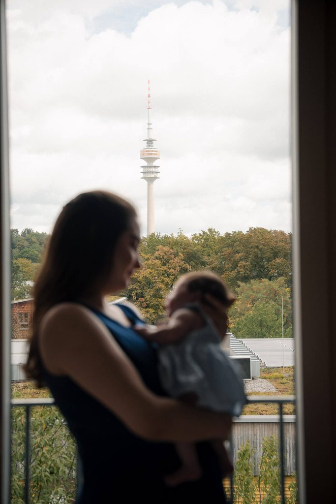 New mom holding her baby at the same window with the Olympiaturm view, weeks after her maternity session